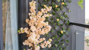 Un árbol en maceta con flores rosas, un muro con celosía y plantas trepadoras, y un balcón con césped artificial verde.