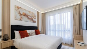 Una habitación de hotel moderna con una cama grande, un televisor de pantalla plana, una obra de arte en la pared y un ventanal con cortinas translúcidas.