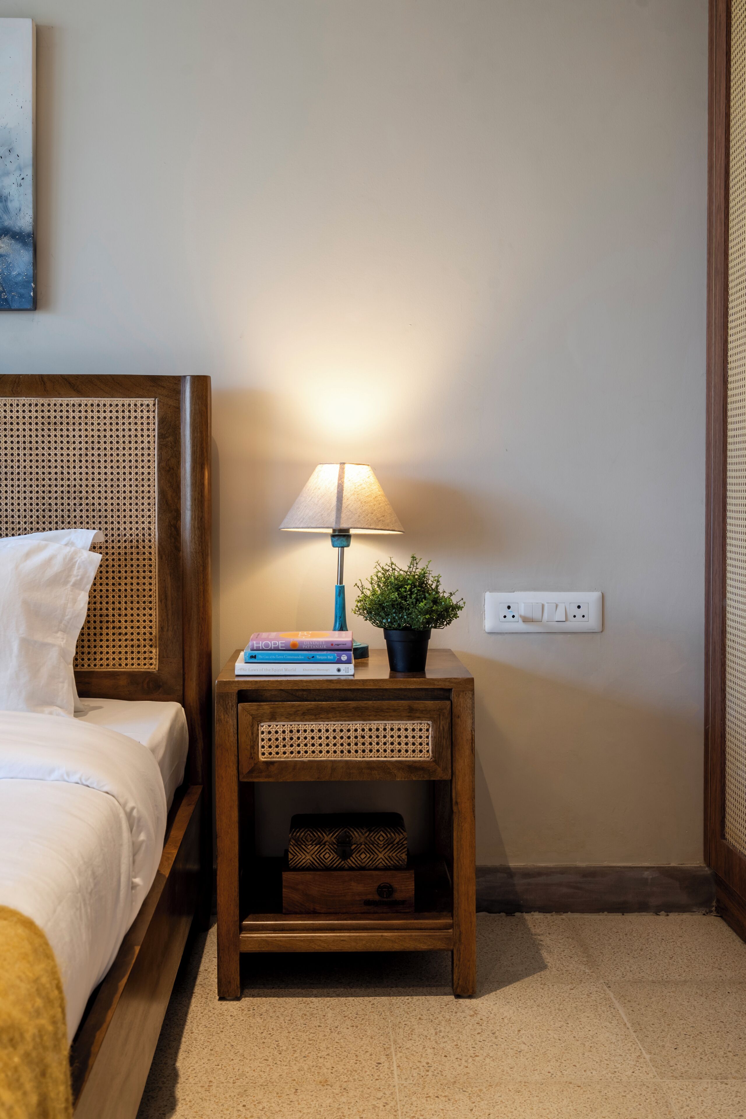 A neatly made bed with a wooden headboard, a bedside table with a lamp, a small plant, and a stack of books.
