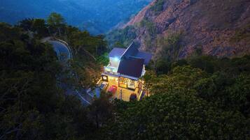 Una casa con techo inclinado, rodeada de un bosque frondoso y un camino sinuoso cerca.