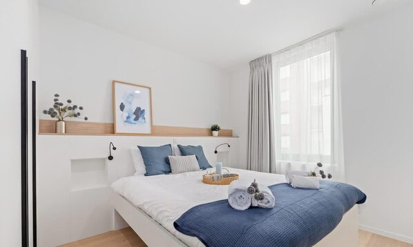 Un dormitorio moderno con cama, almohadas, mesita de noche, un cuadro enmarcado y ventana con cortinas.