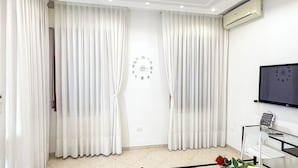 Ein modernes Wohnzimmer mit weißen Vorhängen, einer Wanduhr, einer Klimaanlage, einem Flachbildfernseher und einem Glastisch mit einer Rose.
