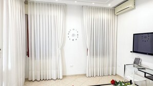 Un salon moderne avec des rideaux blancs, une horloge murale, un climatiseur, un téléviseur à écran plat et une table basse en verre ornée d’une rose.