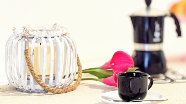 Une lanterne en verre transparent avec une anse en corde, une cafetière noire et une fleur rose sur une table.