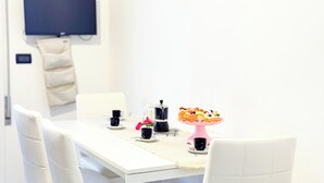 Une table à manger blanche avec des chaises, une machine à café, un présentoir à gâteaux rose avec des cupcakes, et un téléviseur fixé au mur.
