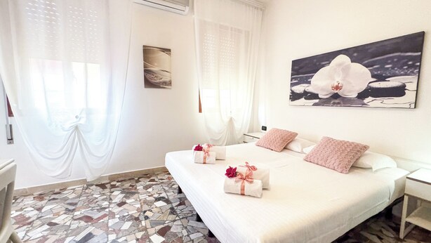 Une chambre d’hôtel avec un grand lit, deux oreillers, une table de chevet, un climatiseur fixé au mur, une fenêtre avec des rideaux légers et un sol en mosaïque.