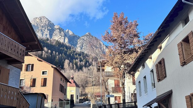 Eine Dorfstraße in den Bergen mit traditioneller Bebauung, einem Schild mit der Aufschrift 'Savilla' und Blick auf die Berge.