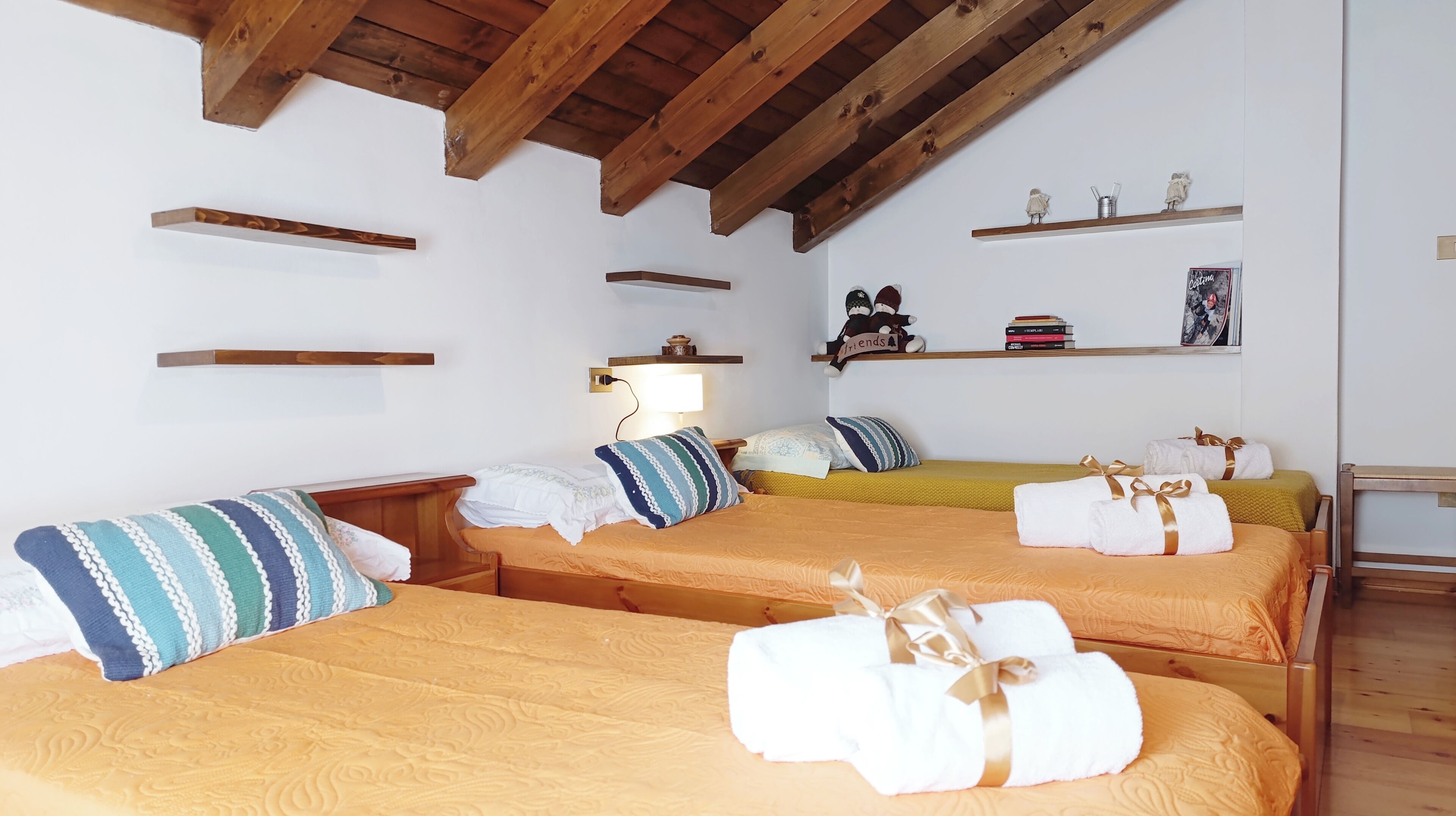 A hotel room with two beds, wooden beams on the ceiling, shelves with books and decorative items, and a bedside lamp.