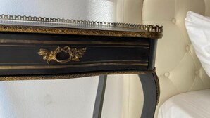 Une table d’appoint noire et dorée, richement décorée, avec un bouton en laiton, placée à côté d’une tête de lit blanche recouverte de tissu.