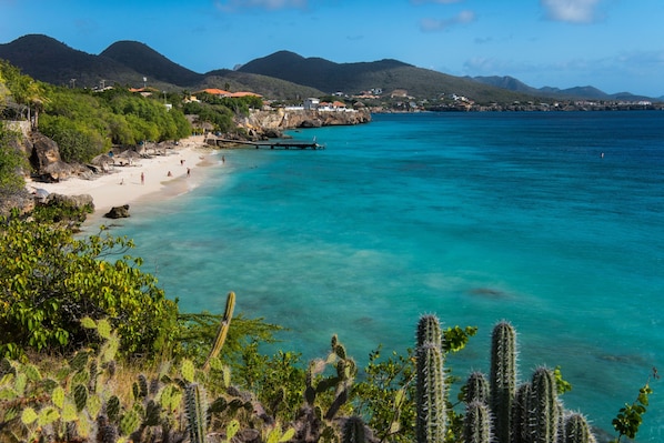 Un paysage côtier caractérisé par une eau turquoise cristalline, une plage de sable et, en arrière-plan, des montagnes.
