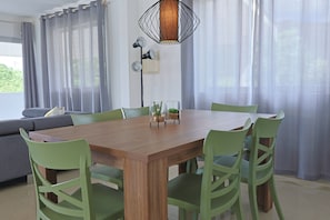 Un coin repas avec une table en bois, des chaises vertes, un luminaire suspendu et des rideaux légers.