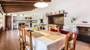 A dining area with a table set for four, wooden chairs, a hanging light fixture, a kitchen with a brick fireplace, and a white wall with shelves. - Villa 'Panada' with Private Pool, Wi-Fi, and Air Conditioning (Pollença)