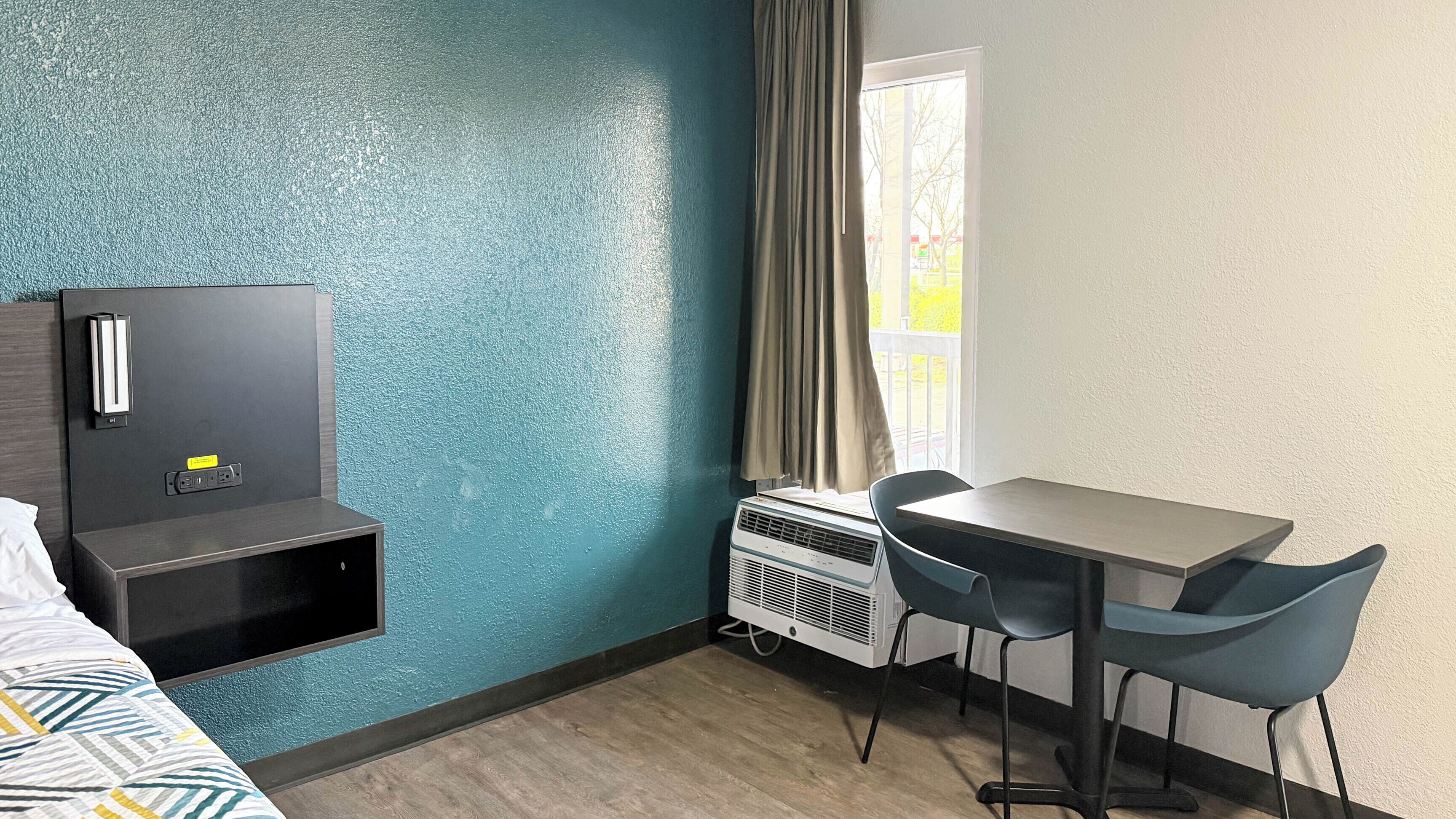 Habitación de hotel con cama, escritorio, dos sillas, aire acondicionado de pared y ventana con cortinas.
