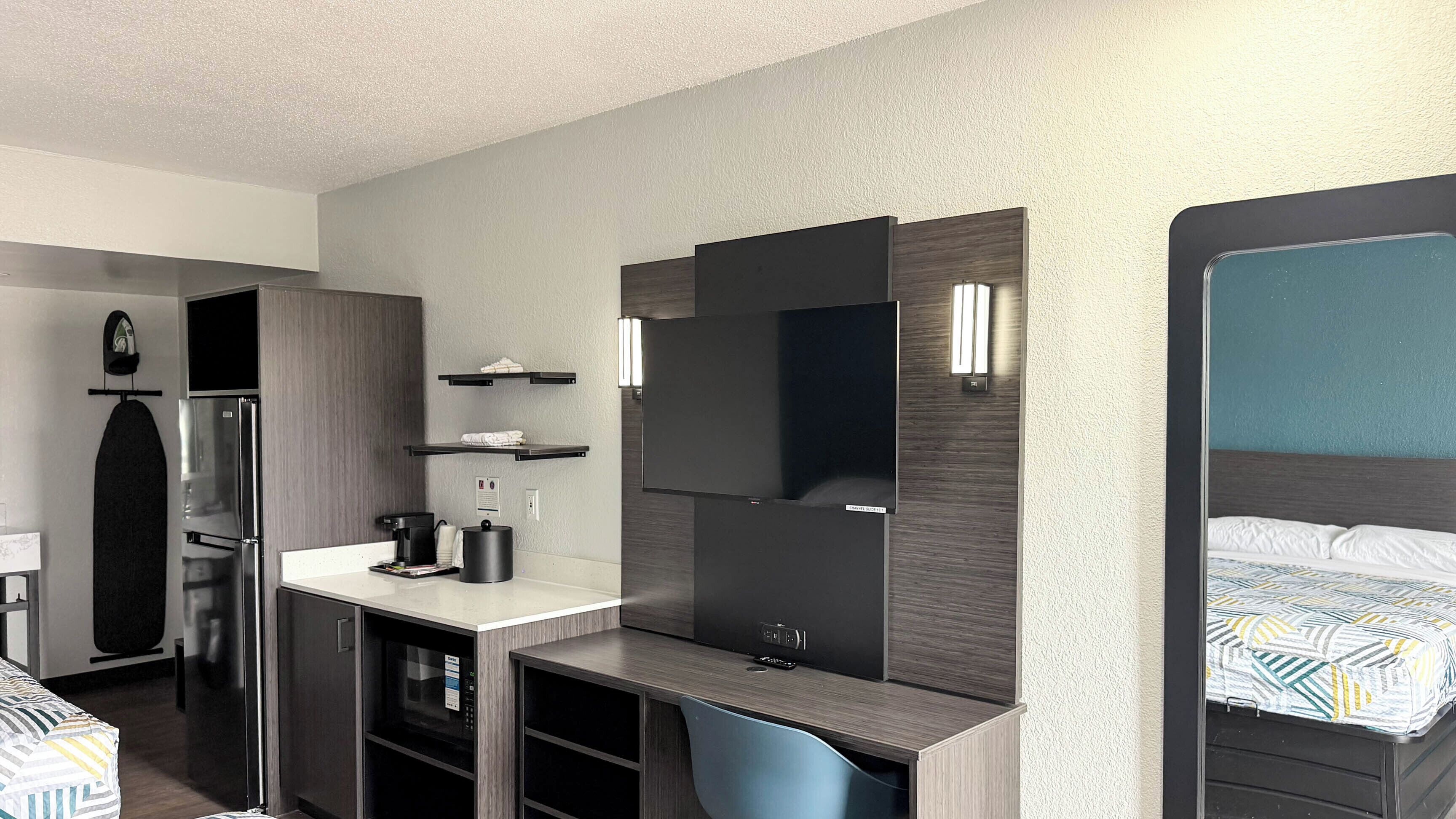 A hotel room with a kitchenette, a bed with patterned bedding, a desk with a chair, and a large mirror.