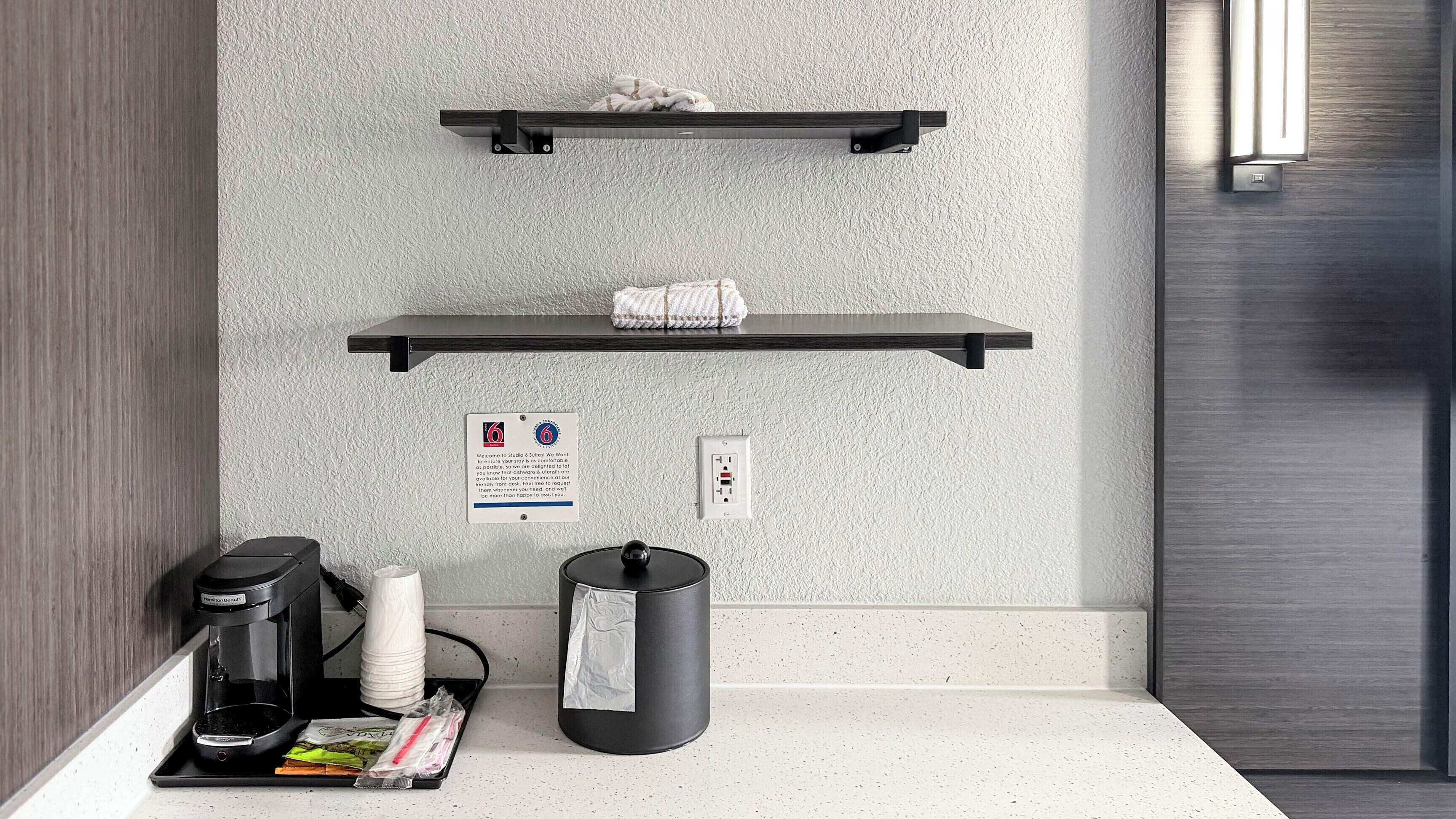 A kitchenette with a coffee maker, a trash can, and a small trash bin on a countertop. There are two black shelves mounted on the wall above the counter.