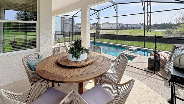 Eine überdachte Terrasse mit einem runden Holztisch, Korbstühlen und Blick auf einen Swimmingpool.