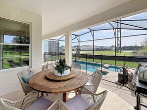 Um alpendre coberto com uma mesa de madeira redonda, cadeiras de vime e vista para uma piscina.