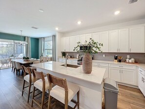 A modern kitchen with a central island, white cabinetry, and a large window providing natural light. - Reunion Retreat: Private Pool/Spa (Kissimmee)