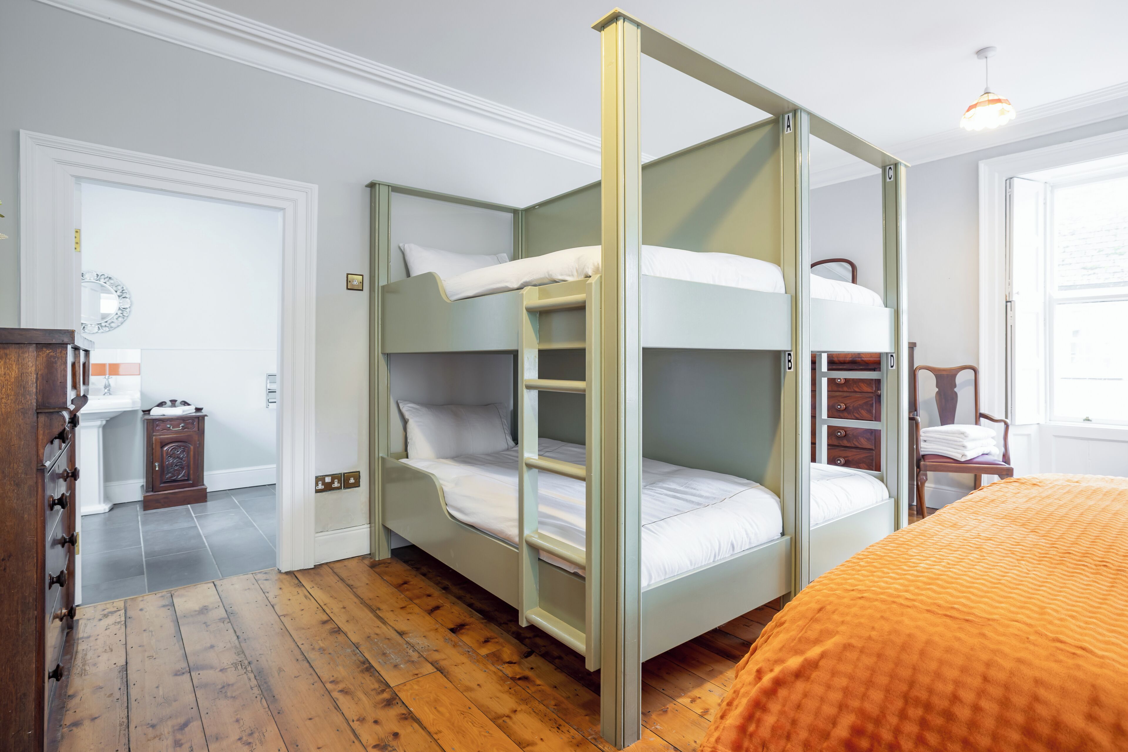 Una cama litera con escalera, un aparador de madera y una silla en una habitación con piso de madera y paredes blancas.