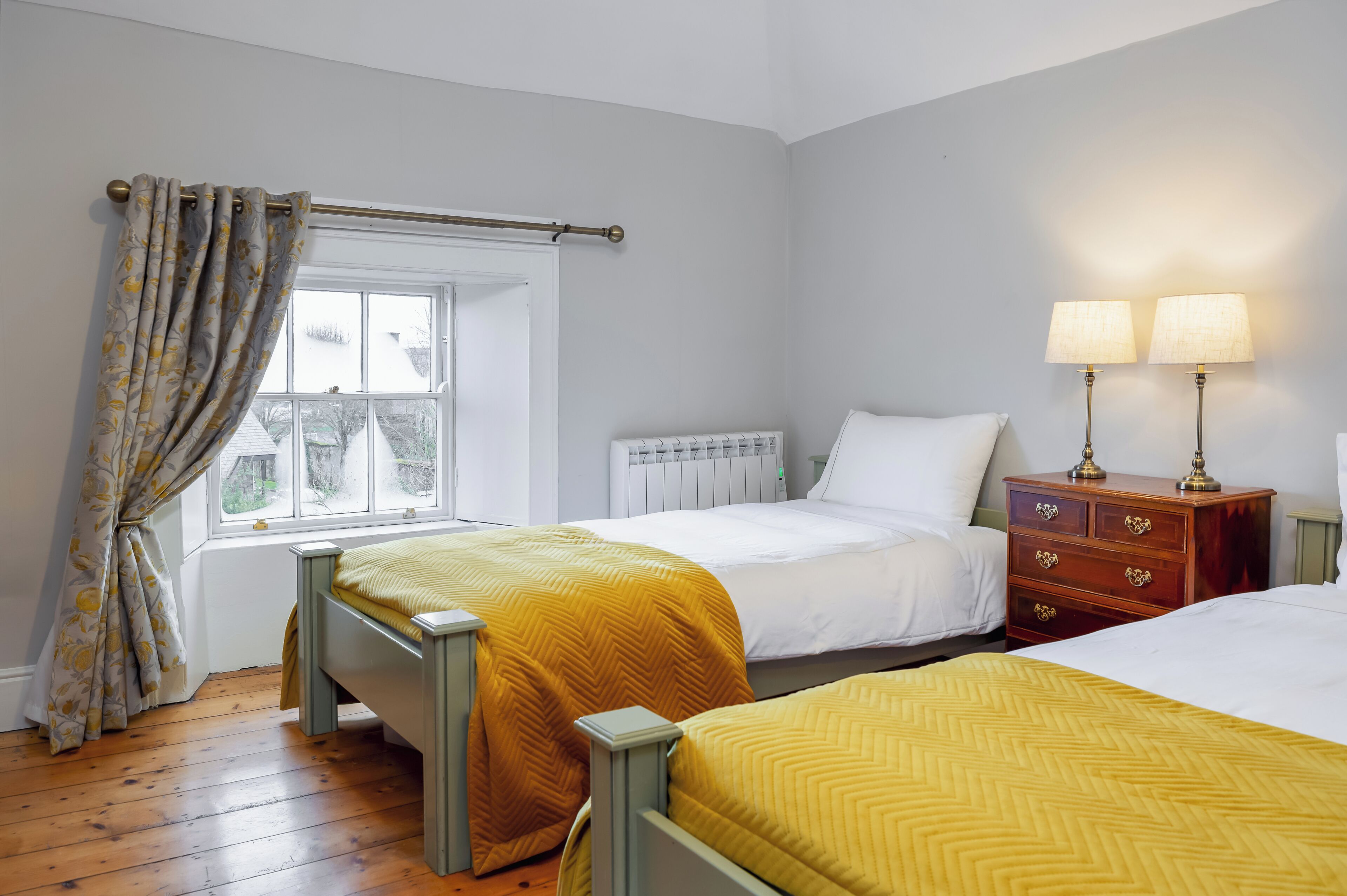 A bedroom with two beds, a window with curtains, a radiator, and a nightstand with lamps.