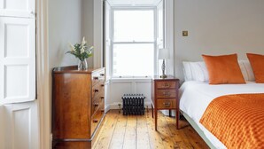 A bedroom with a wooden dresser, a radiator, a window, and a bed with orange pillows.