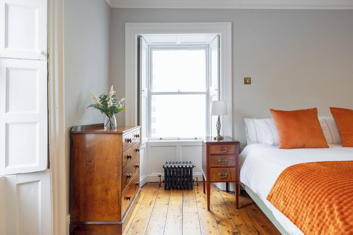 A bedroom with a wooden dresser, a radiator, a window, and a bed with orange pillows.