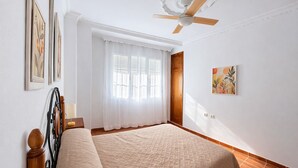 A bedroom with a bed, a ceiling fan, a window with curtains, a door, and a painting on the wall. - Apartamento Arrumbadores con terraza en Conil (conil de la frontera)