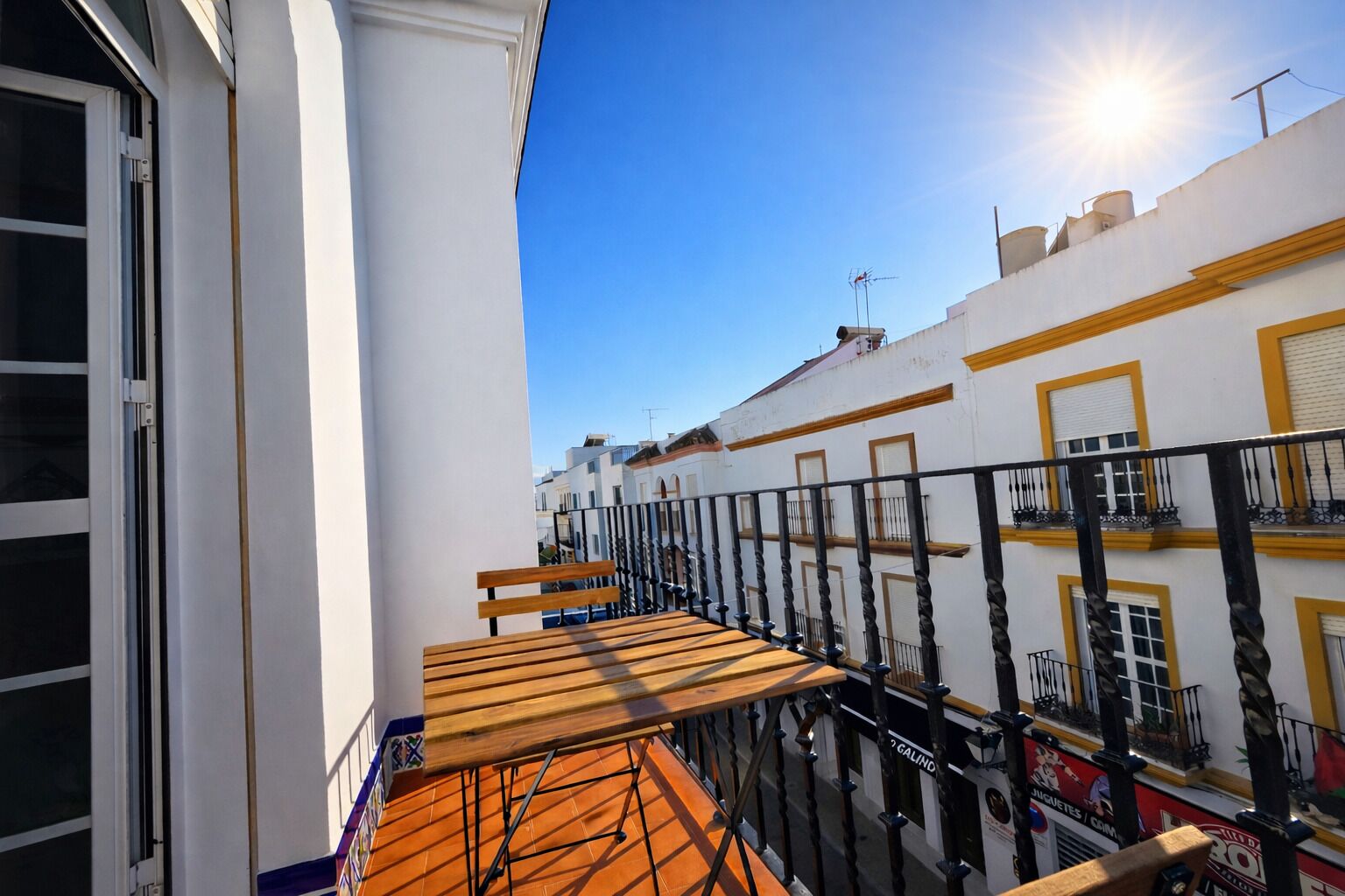Ein Balkon mit einer Holzbank, weiße Gebäude mit gelben Verzierungen und ein klarer, blauer Himmel.