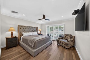 A bedroom with a large bed, two bedside tables with lamps, a ceiling fan, a TV mounted on the wall, and a window with a view.