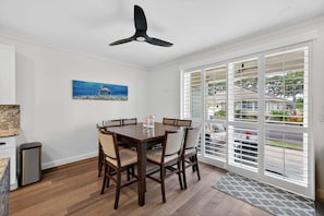 A dining area with a table and chairs, a ceiling fan, a painting, and a view through large windows.