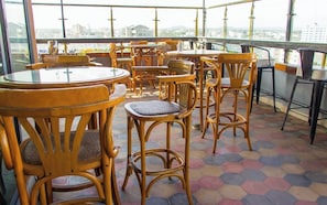 Een dakterras met ronde houten tafels en stoelen, een betegelde vloer en uitzicht op de stad.