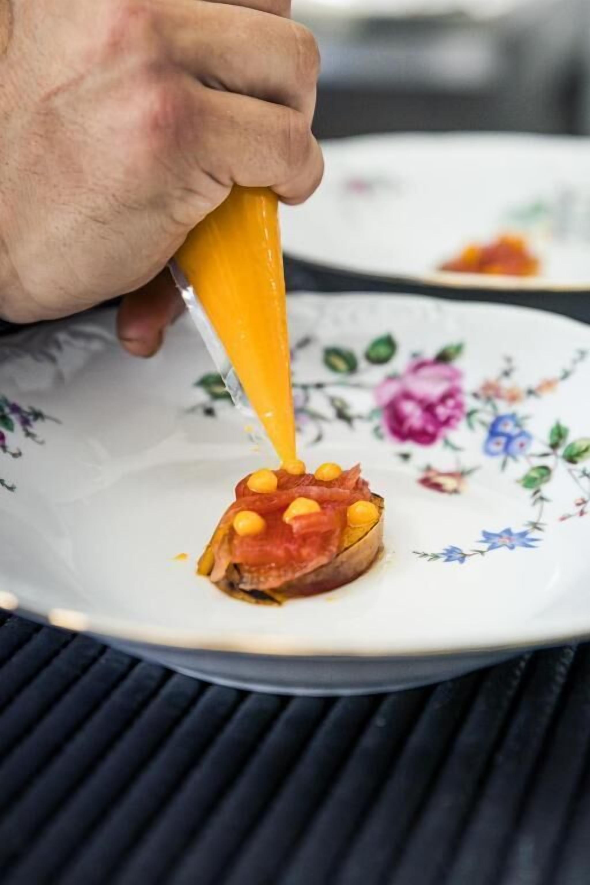 Una mano sostiene un envase exprimible, probablemente con un condimento, aplicándolo sobre un alimento en un plato.
