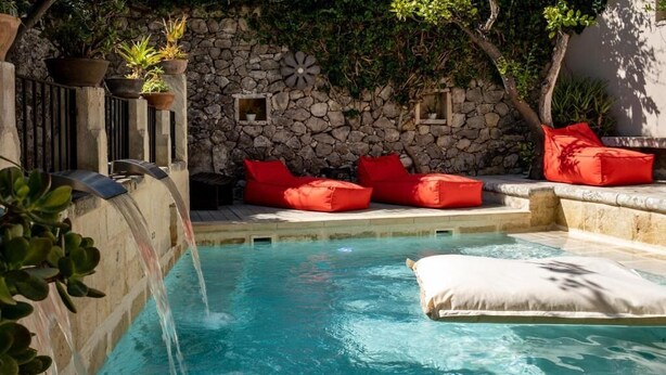 Une piscine avec un mur en pierre, des coussins rouges et une cascade décorative.