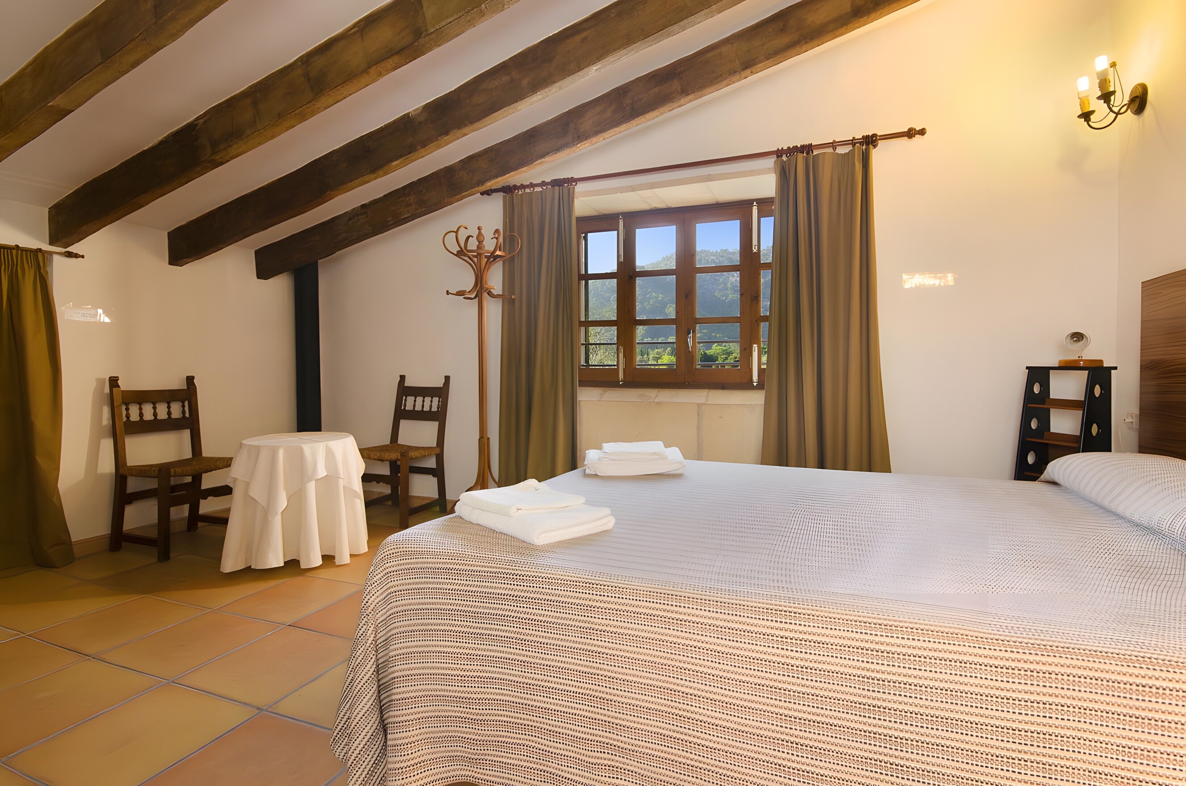 A bedroom with a large bed, a small round table, two chairs, a window with curtains, and a wooden ceiling beam.