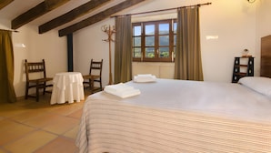A bedroom with a large bed, a small round table, two chairs, a window with curtains, and a wooden ceiling beam. - Holiday Home "Camp Redo" with Mountain View, Pool, Garden, Terrace & Wi-Fi (Illes Balears)