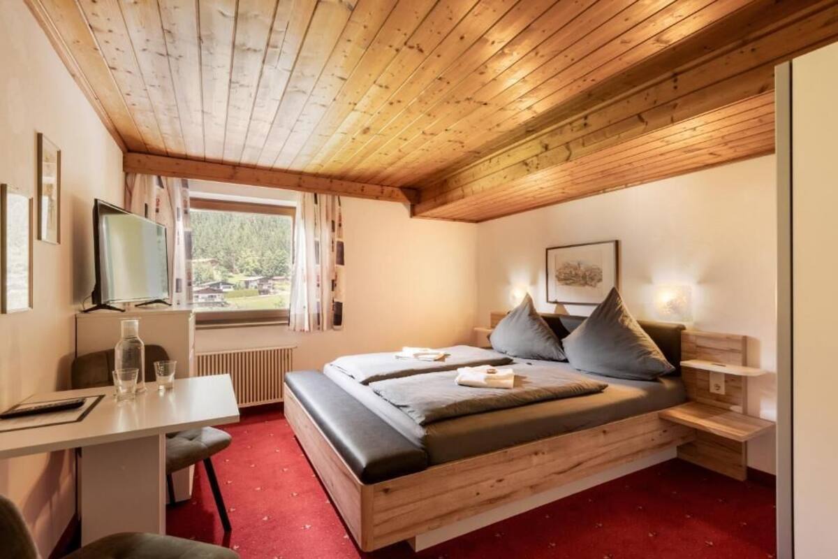 Ein Schlafzimmer mit Holzboden, einem Bett mit grauer Bettwäsche, einem kleinen Tisch, einem Stuhl, einem Fernseher und einem Fenster mit Vorhängen.