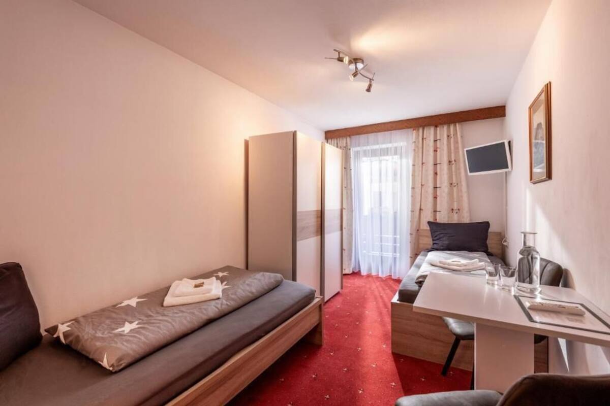 Ein kleines Hotelzimmer mit einem Einzelbett, einem Schreibtisch mit Stuhl, einem Fernseher und einem Fenster mit Vorhängen.
