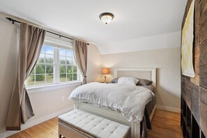 A bedroom with a bed, a bench, a window with curtains, and a lamp. - Wharfside Watch – NEW Oceanfront Home! (Blue Hill)