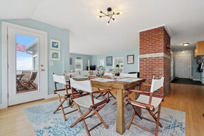 A dining area with a wooden table and chairs, a brick column, and a view through a door to an outdoor patio. - Wharfside Watch – NEW Oceanfront Home! (Blue Hill)