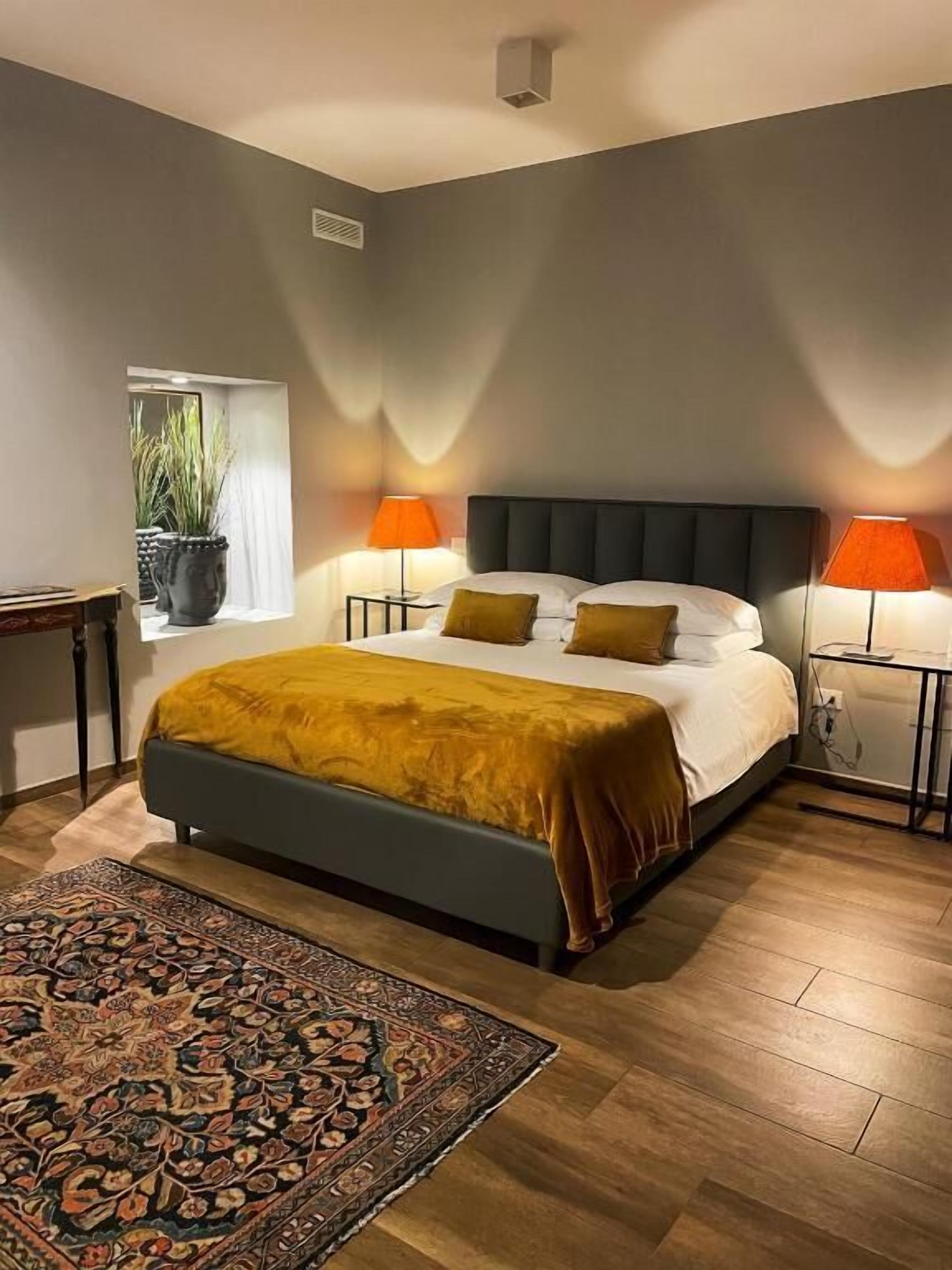 A bedroom with a bed, two bedside lamps, a nightstand, a potted plant, and a patterned rug.