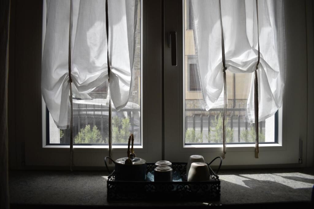 Una habitación con dos ventanas, cada una con cortinas blancas, una mesita con tetera, tazas y un bandeja.