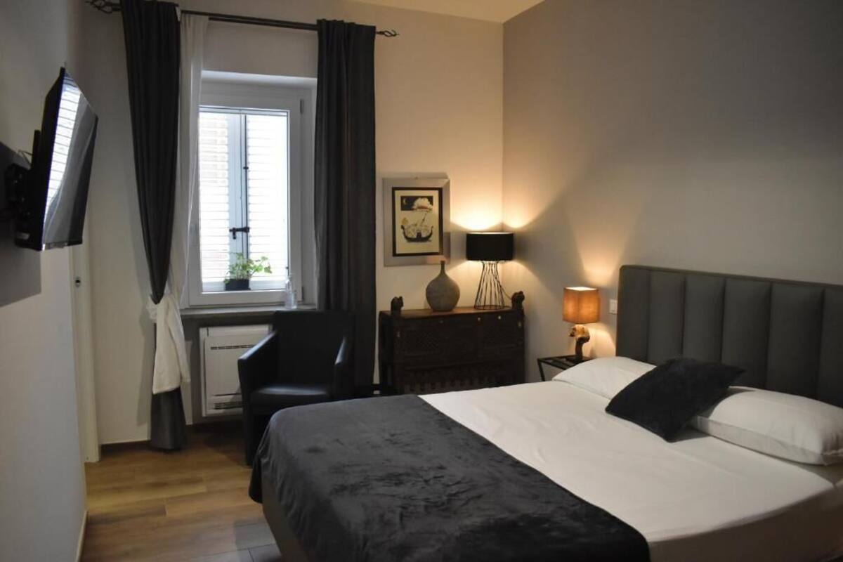 A bedroom with a bed, a desk, a chair, a window with curtains, a wall-mounted TV, and a bedside lamp.
