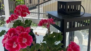 Un balcón con geranios en macetas, un farol negro y vistas a una colina con edificios.