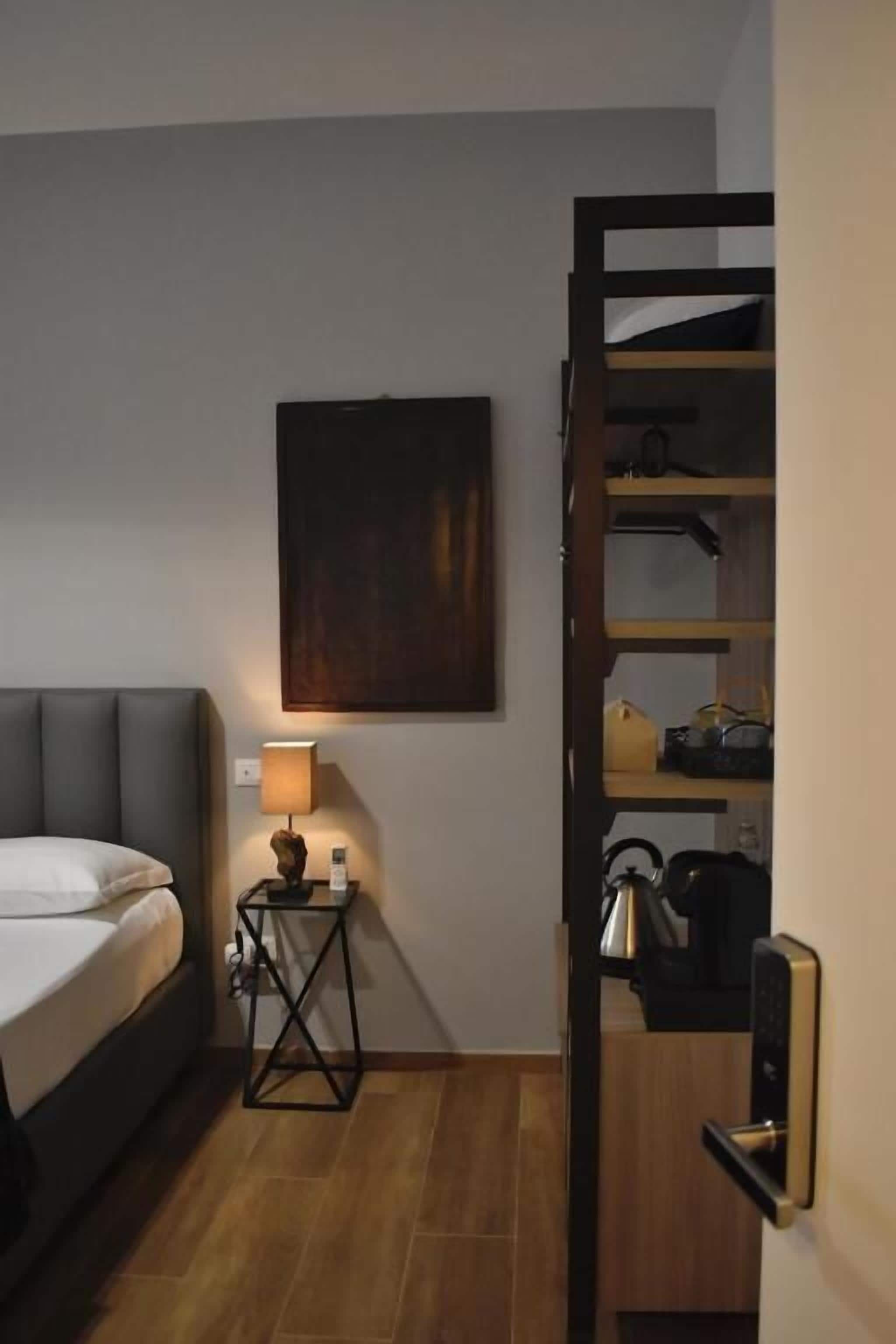 Une chambre d’hôtel comprenant un lit, une table de chevet avec une lampe, un tableau accroché au mur, un plancher en bois et une étagère intégrée contenant des ustensiles de cuisine.