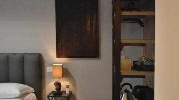 Une chambre d’hôtel comprenant un lit, une table de chevet avec une lampe, un tableau accroché au mur, un plancher en bois et une étagère intégrée contenant des ustensiles de cuisine.
