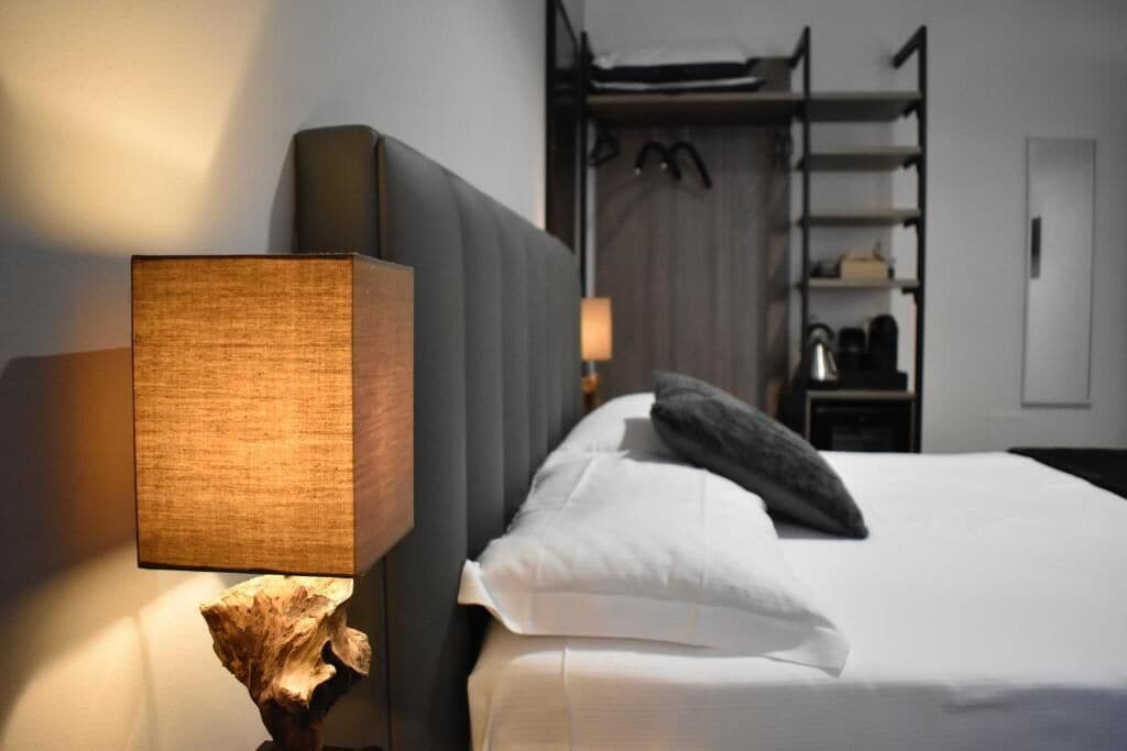 A hotel room with a bed, a nightstand with a lamp, and a wardrobe in the background.