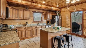 A wooden cabin kitchen with granite countertops, wooden cabinets, a central island with stools, and a large window overlooking a deck. - Spacious*Private Pool*Home Theater*Close to Pigeon Forge (PIGEON FORGE)