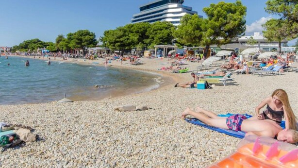 A pebbled beach with people sunbathing, a building in the background, and trees. - Apartments Milka Vodice (Vodice)