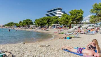 A pebbled beach with people sunbathing, a building in the background, and trees.