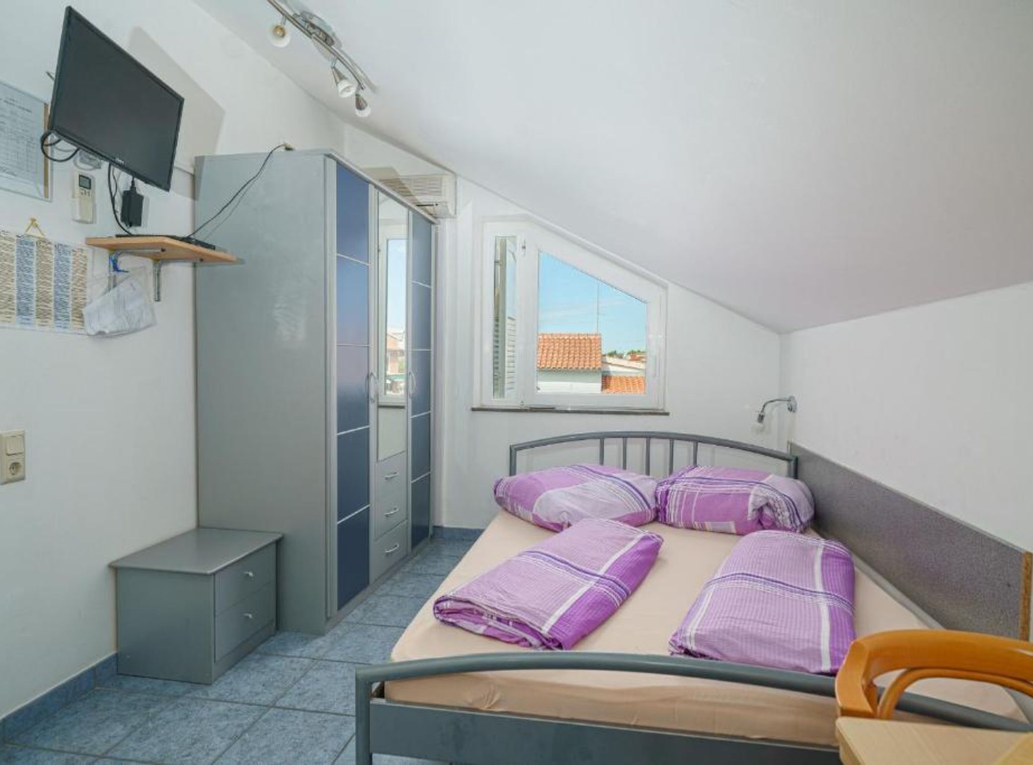 Une petite pièce propre et bien rangée, avec un lit, un bureau, une chaise, une armoire, un téléviseur et une fenêtre donnant sur des immeubles.